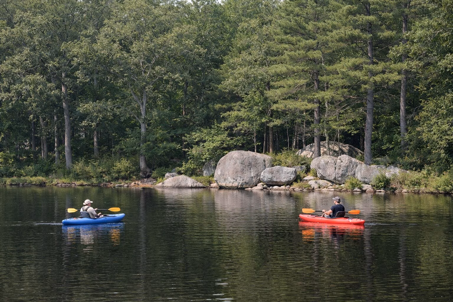 Two kayakers paddling a calm pond below a wooded shore with boulders