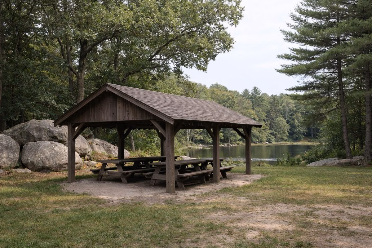 Open-sided wooden picnic shelter in a grassy clearing near a pond